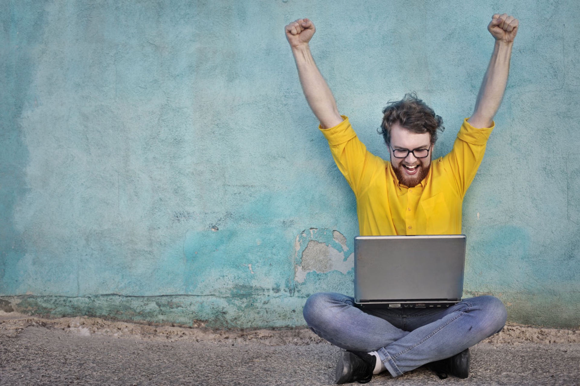 Ein Mann in gelbem Pullover sitzt im Schneidersitz mit dem Laptop auf dem Schoß und ausgestreckten Armen und geballten Fäusten in der Luft.