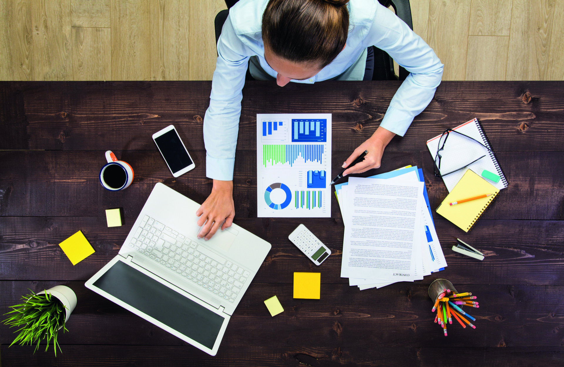 Draufblick auf eine Frau, die am Schreibtisch mit Planungsunterlagen sitzt und auf Ihrem Laptop tippt.